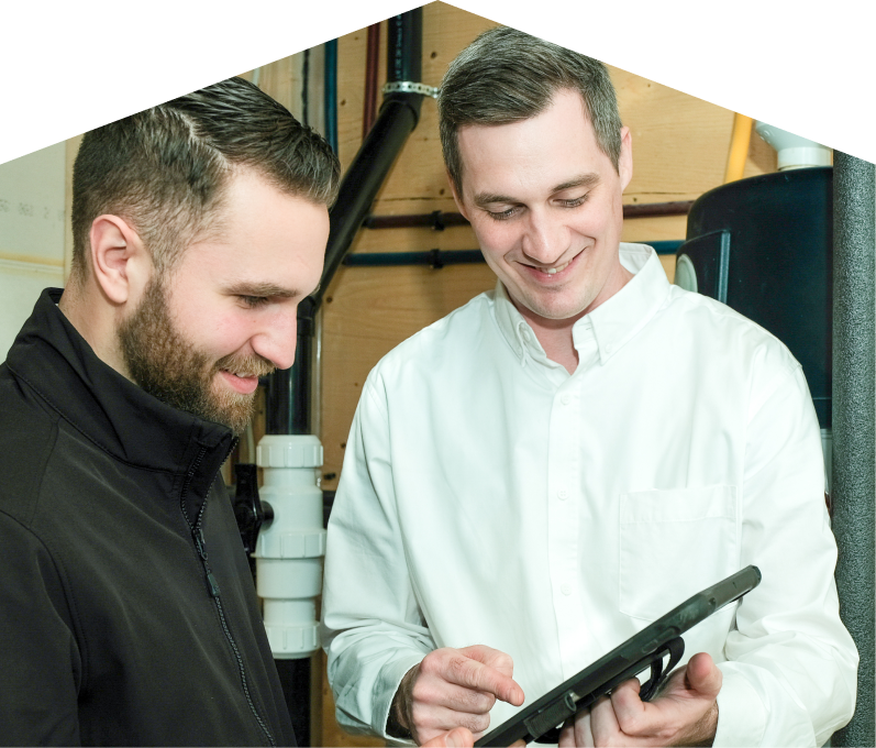 669 Heat specialist reviewing maintenance plan options with a smiling homeowner using a tablet, inside a utility room with visible piping. 669 Heat specialist reviewing maintenance plan options with a smiling homeowner using a tablet, inside a utility room with visible piping.