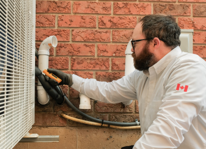 A 669 Heat HVAC specialist wearing glasses and gloves uses a diagnostic tool to service an outdoor AC unit beside a brick wall. A 669 Heat HVAC specialist wearing glasses and gloves uses a diagnostic tool to service an outdoor AC unit beside a brick wall.